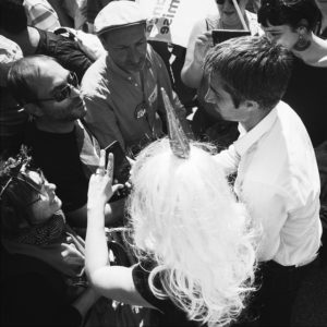 A black and white image of a group of people engaged in conversation at a fête. The center of attention is a person in a white shirt talking with someone wearing a long, white wig and a unicorn horn. Other people around them are holding phones, listening, and observing.