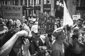A black and white photo of a crowded street protest. People hold various signs, including one that says "MACRON PLOUTOCRATE" and another with various figures and facts. The atmosphere is intense as protesters make their voices heard amidst buildings, giving the scene an almost fête-like fervor.