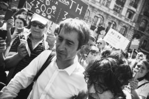 A black and white photograph captures a group of people at a protest or rally. One person in the foreground holds a sign with various symbols, while another sign in the background reads "fête à Macron" in French. The individuals are using their phones to take pictures or videos.
