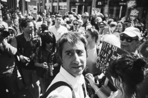 A grayscale photo captures a man in the foreground amidst a bustling crowd during the La fête à Macron protest. People around him hold various signs and banners, while others take photos or engage in conversation. The scene is lively, with city buildings visible in the background.