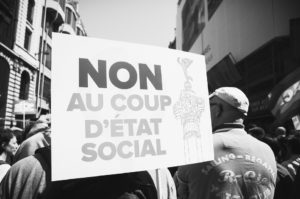 A black-and-white photo shows a group of people at a protest. One person in the foreground holds a sign reading "Non au coup d'état social" with an image of a column. Other protesters and buildings can be seen in the background, highlighting the passion and energy reminiscent of La fête à Macron.