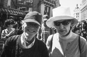 A black and white photo shows an older man and woman standing closely together on a bustling city street during La fête à Macron. The man is smiling, wearing a baseball cap and sweater, while the woman sports a sun hat, sunglasses, and jacket. Both carry backpacks amidst the lively backdrop of people and buildings.