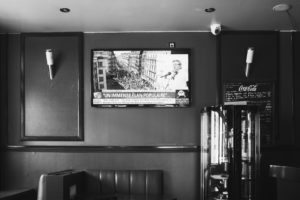 Black and white photo of a room with wall-mounted light fixtures. A television above shows a news broadcast covering La fête à Macron with an image of a man speaking and a crowd below. A cabinet with glass doors stands to the side, and a small Coca-Cola menu is visible on the wall.