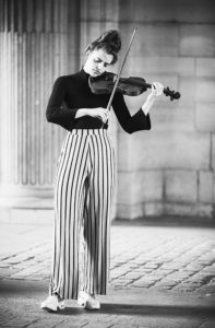 A woman debout, wearing a black turtleneck and striped wide-leg pants, stands on a cobblestone surface, joue the violon with a focused expression. The black and white photo is taken outdoors, with large decorative pillars in the background.