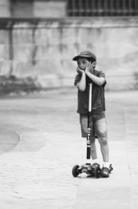 Le petit Oscar stands next to their scooter on a cobblestone street. The child is wearing a cap, a t-shirt, shorts, and sandals, with their hands resting on their face, looking thoughtful or bored. The image is in black and white.