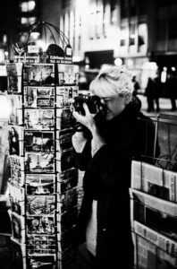 Black and white photo of Maud with light-colored hair taking a picture with a camera. She's standing next to a rotating postcard stand filled with postcards of Paris, à l'affut of the perfect shot. The background is blurred, indicating a nighttime urban setting with bright lights.