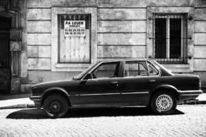 Black and white photo of a parked sedan in front of an old building with barred windows. A sign in French on the building reads "À Louer" with contact phone numbers. The scene appears to be on a quiet, cobblestone street, evoking an atmosphere à l'ancienne.