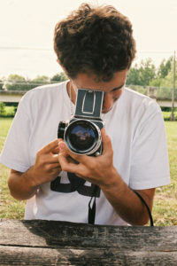 A person with curly hair is seated at a wooden table outdoors, holding a Hasselblad camera at eye level, looking through the viewfinder. The person is wearing a white t-shirt, and the background shows greenery and a bridge in the distance, capturing the essence of Grégoire's passion for photographie.