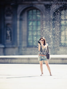 Jenny Star, wearing sunglasses, a sleeveless top, and shorts, stands outdoors capturing a photo with her camera. She has a shoulder bag. The background includes a building with large windows and a blurred fountain with water droplets visible.