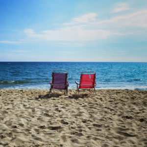 Two empty beach chairs, one red and one purple, sit cote à cote on the sandy shore facing the calm blue ocean under a partly cloudy sky, depicting a serene and peaceful seaside scene.