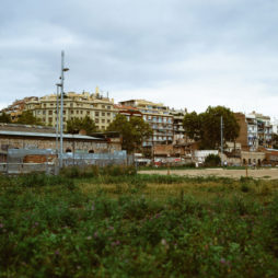 Overcast cityscape in Barcelona featuring a mix of modern and older buildings. In the foreground, a grassy area with a few trees surrounds an abandoned lot. The buildings in the background are of varying heights, some with graffiti, and one showcasing a prominent graffiti-covered fence.