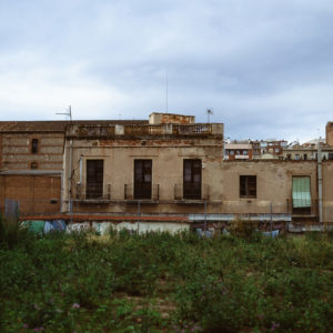 An old, abandoned building with a weathered, beige exterior and barred windows stands behind a barbed wire fence in the outskirts of Barcelona. The foreground features overgrown grass and wild plants, reminiscent of the Jardi Comestible in Catalonia, while urban buildings rise under a cloudy sky.