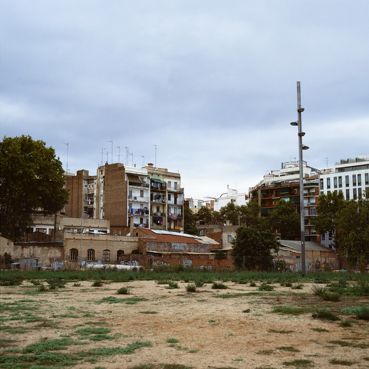 An urban scene in Barcelona featuring a vacant, grassy lot with patches of dry earth in the foreground. In the background are several multi-story residential buildings with balconies, some with laundry hanging. The overcast sky looms above, and a tall streetlight stands on the right, capturing Catalonia's essence.