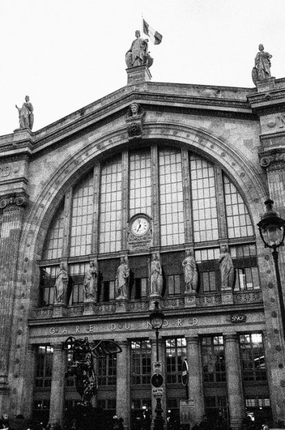 A grayscale image of the Gare du Nord Paris train station. The ornate facade features large arched windows, statues on the roof, and a central clock. The French flag flies at the top. Pedestrians and a few lampposts are visible in the foreground.