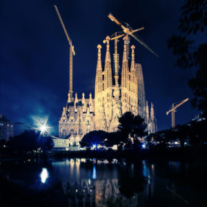 Night view of La Sagrada Família, an illuminated basilica in Barcelona under construction. Tall cranes surround its intricate spires, and the building's reflection shimmers in a nearby pond. The dark sky contrasts with Sagrada Familia's bright facade.