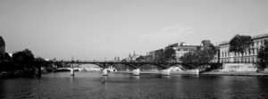 A black-and-white photograph of a serene river with multiple bridges, including the Pont des Arts, spanning across it. Historic buildings and trees line the riverbanks under a clear autumn sky.