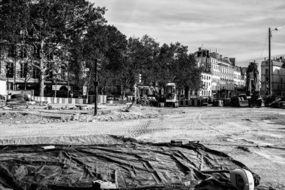 A black and white photo depicting a large construction site at Place de la Bastille in an urban area. Heavy machinery and construction equipment are scattered across the dirt terrain, indicative of ongoing renovation efforts. Buildings and trees are visible in the background, with a streetlight at the left edge of the image.