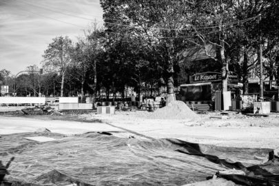 A black-and-white image showing a street under construction near Place de la Bastille. Several trees line the road, and a pile of dirt is visible on the ground. A building in the background displays a sign that reads "Le Kiosque." The area appears to be fenced off for ongoing rénovation work.