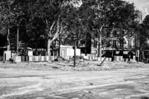 A black-and-white image of a construction site on a street near Place de la Bastille, surrounded by trees and buildings. Various construction materials and fenced-off areas are visible. The area appears to be busy with Rénovation activities, as some workers and vehicles can be seen in the background.