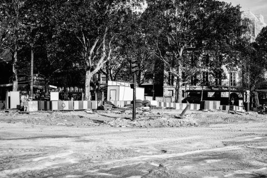 A black-and-white image of a construction site on a street near Place de la Bastille, surrounded by trees and buildings. Various construction materials and fenced-off areas are visible. The area appears to be busy with Rénovation activities, as some workers and vehicles can be seen in the background.