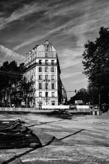 Black and white image of a city street undergoing rénovation. The focus is on a tall, narrow building with multiple windows and a slanted roof. The site is surrounded by barriers and rubble, with trees and power lines visible near place de la Bastille. The sky is lightly clouded.