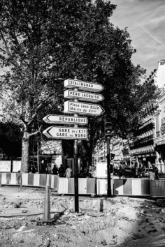 A black and white photo depicts street signs in Paris pointing towards Stalingrad, Père Lachaise, Place Léon Blum-Mairie du 11éme, République, and Gare de l'Est-Gare du Nord. The background shows trees, buildings under rénovation, construction barriers, and pedestrians near place de la Bastille.