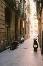 Narrow alleyway in Barcelone's Barri Gotic, flanked by old buildings with balconies. Several scooters are parked along one side, with sunlight illuminating the street. Faint graffiti is visible on one of the closed shop doors. The ambiance is calm and quiet.