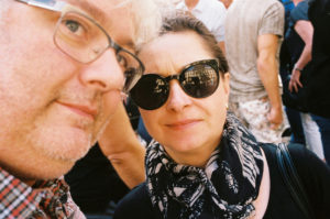 Close-up of a man and a woman, with the man wearing glasses and partially visible on the left and the woman wearing dark, round sunglasses and a patterned scarf on the right. Both are looking at the camera with an air of mystery in this "Sans titre" shot, as a crowd of people blurs in the background.