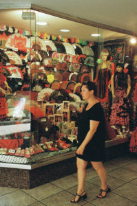 A woman in a black dress walks past a magasin display full of colorful fans, flamenco dresses, and accessories. The window is packed with various traditional Spanish items, including decorative fans, polka dot shoes, and framed pictures.