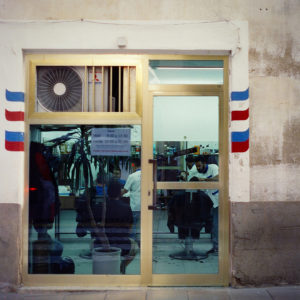 A barbershop viewed from outside through a glass door and window, with gold frames. The entrance is flanked by blue, white, and red stripes. Inside, barbers are cutting clients' hair amid various equipment, creating a busy scene worthy of a "Sans titre" masterpiece.