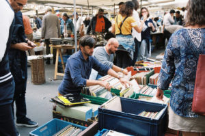 A crowded outdoor market where people are browsing through vinyl records in boxes. A man on the left in a navy shirt is looking at records, while others around him, including a woman in a blue patterned shirt, are also searching through the bins for that perfect find—la bonne pioche.