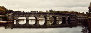 A panoramic view of an ancient stone bridge with multiple arches reflecting on a calm river in Le Cosson. The bridge is surrounded by the autumn foliage of Domaine de Chambord, with trees displaying a mix of green, yellow, and orange leaves. The overcast sky casts a subdued light on the scene.