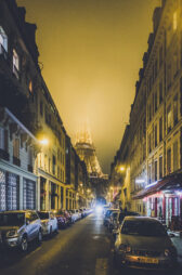 A narrow street in Paris at night with cars parked on both sides, leading towards the illuminated Eiffel Tower shrouded in brume. The street is lined with tall buildings with glowing windows and streetlights, adding a warm lumière to the scene.