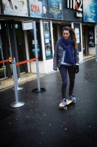 A woman with a blue scarf and gray jacket rides a skateboard on a wet sidewalk in front of a theater with movie posters and barriers near the entrance.