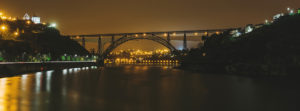 A panoramic night view of the lit-up Pont São João in Porto, Portugal, spanning over a wide river. The bridge has an arch structure, and bright lights illuminate the water's reflection. Buildings and lights are visible on both sides of the riverbank, creating a serene urban nightscape.