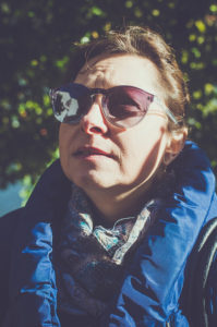 A belle woman with short hair stands outdoors, wearing sunglasses and a blue winter coat. Sunlight casts a shadow on her face as she looks into the distance. The background is blurred with green foliage, adding to the serene atmosphere of the ville.