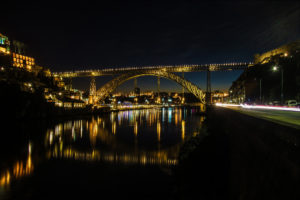 A night view of the Dom Luís I Bridge in Porto, Portugal, illuminated with golden lights. The bridge spans across the Douro River, reflecting its lights on the calm water below. Buildings on both sides are also lit, adding to the picturesque scene.