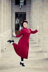 A person dressed in a red vintage coat and hat with floral accents joyfully poses like Mary Poppins, with one leg kicked up, arms bent, and hands pointing outward. The background features an elegant colonnade with tall pillars and a distant doorway.