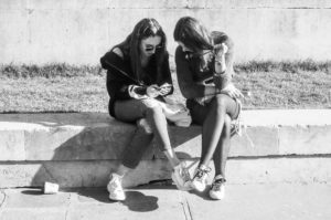 Camille et Noémie sit on a low concrete wall outdoors, closely looking at something on a phone that one of them is holding. Both wear casual clothing, including jackets and sneakers. The setting appears sunny with grass behind them and shadows on the pavement.