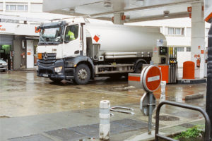 A white camion with a silver citerne is parked at a gas station near a fuel pump. The truck has a hazard placard, and the area is wet, with caution signs and station equipment visible, including a red stop sign for restricted access.
