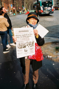 A person wearing a hat and glasses stands on a city street holding up a French newspaper with the headline "Macron voudrait en finir avec les Gilets jaunes... Mais ils sont toujours là". A bus is visible in the background, and other people are always there on the bustling street.