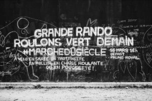 A black and white wall with chalk writings in French, promoting a march event. The main text reads, "Grande Rando Roulons Vert Demain #Marchedusiècle 16 Mars 16H Départ Palais Royal." Surrounding graffiti includes other messages, drawings, and doodles in the heart of Paris.