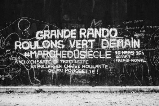 A black and white wall with chalk writings in French, promoting a march event. The main text reads, "Grande Rando Roulons Vert Demain #Marchedusiècle 16 Mars 16H Départ Palais Royal." Surrounding graffiti includes other messages, drawings, and doodles in the heart of Paris.