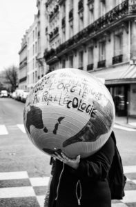 A black-and-white photo features a person standing on a street during the Marche du siècle in Paris, holding a large, inflated globe that obscures their head and upper body. The globe has handwritten French text mentioning ecology and climate change. The background includes buildings and parked cars.