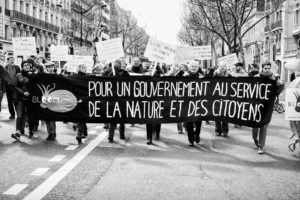 Black and white photo of a group of protesters marching down a street in Paris, holding a large banner that reads "Pour un gouvernement au service de la nature et des citoyens" (For a government serving nature and citizens). Other demonstrators hold various signs during the iconic Marche du siècle.