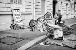 A person wearing a coat is seen adjusting a large, fallen paper-mâché statue resembling the Statue of Liberty on a Paris sidewalk. The statue holds a sign reading "Plan Anticor Très Urgent." Another person walks away in the background on the street, hinting at the vibrant life of France's capital.