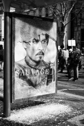 Black and white photo of a damaged Sauvage bus stop advertisement featuring a stern-looking man with facial hair and a tattoo. The glass of the ad is broken, lying shattered on the ground. People are marching in the background, holding up signs.