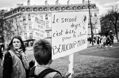 A black-and-white photo captures people at a protest in Paris. One person in the foreground holds a sign in French that reads, "Le second degré, c'est drôle, pour le climat beaucoup moins." Another sign in the background mentions "pollution." This scene from the Marche du siècle unfolds in an urban area.