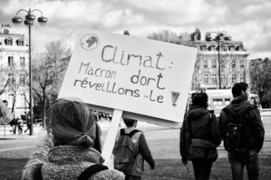 A grayscale photo shows a person in Paris holding a sign that reads, "Climat: Macron dort réveillons-le!" at the Marche du siècle. Other individuals walk in the background, with historic buildings and a cloudy sky visible. The sign is a protest call concerning climate change.