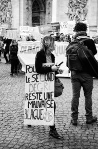 A black-and-white photo shows a woman at a protest rally in Paris on 16 Mars 2019. She is wearing a sign around her neck that reads, "Pour que le second degré reste une mauvaise blague," translated to, "So that satire remains only a bad joke." Other protesters from the Marche du siècle with banners are in the background.